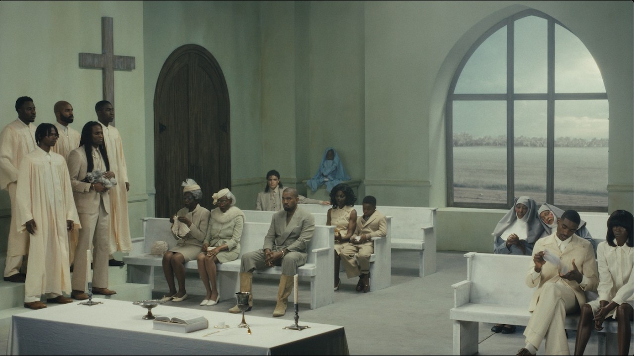 Cena do videoclipe Father de Kanye West, dirigido por Bianca Censori, com o rapper sentado em uma igreja cercado de personagens surreais